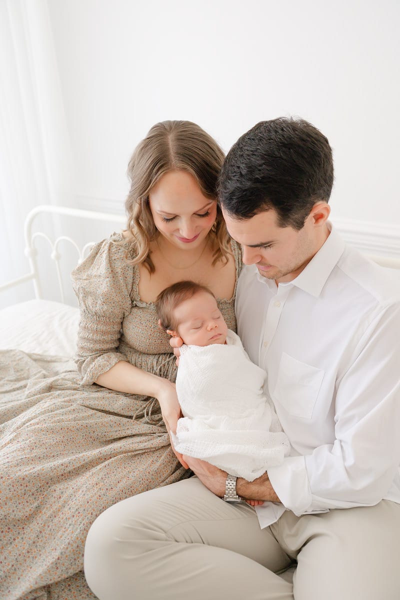 Parents sitting together on a white iron bed gazing at their sleeping newborn during an Emerald Coast newborn photography session