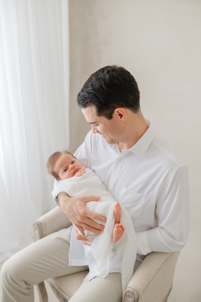 Dad holding his alert newborn in a chair during an Emerald Coast newborn photography session in Niceville Florida