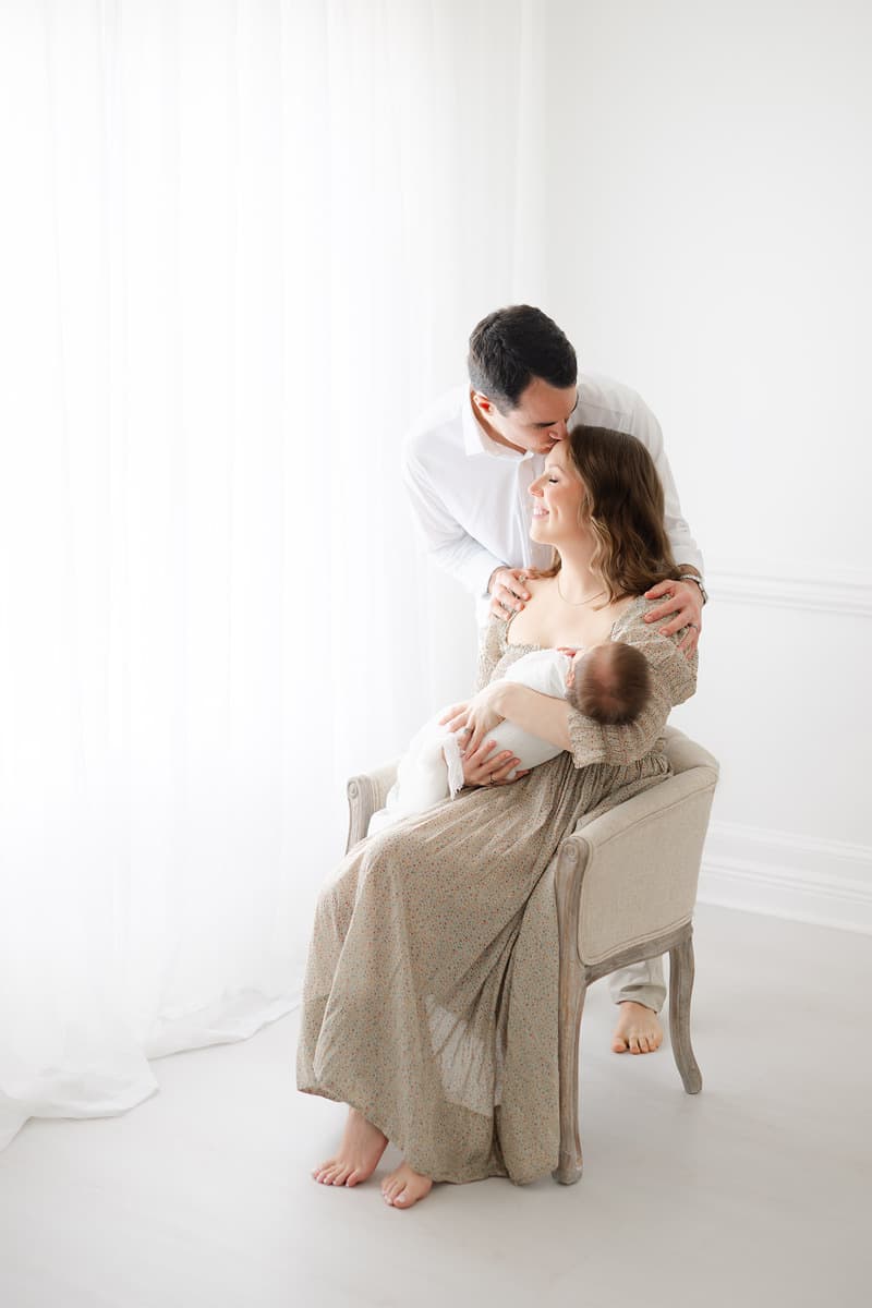 Emerald Coast newborn photographer captures dad kissing mom while she holds their sleeping newborn in a bright airy studio