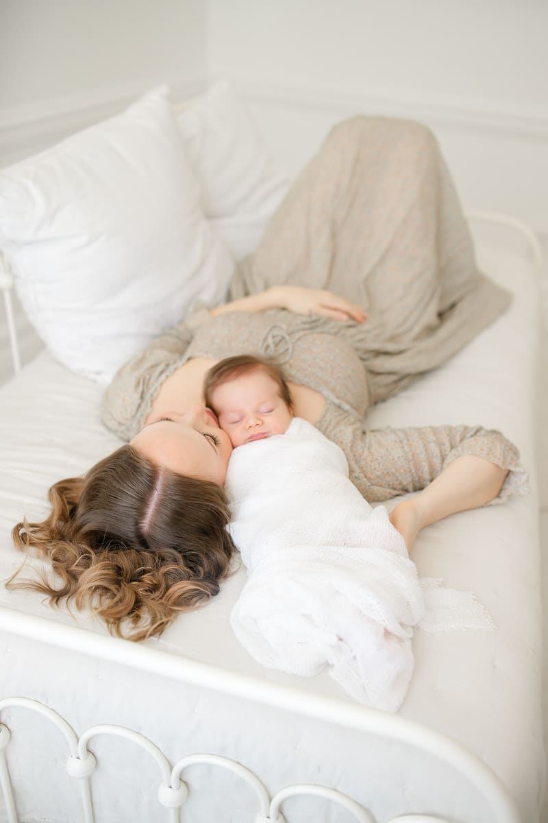Mom lying beside her sleeping newborn on a white bed during a lifestyle Emerald Coast newborn photography session
