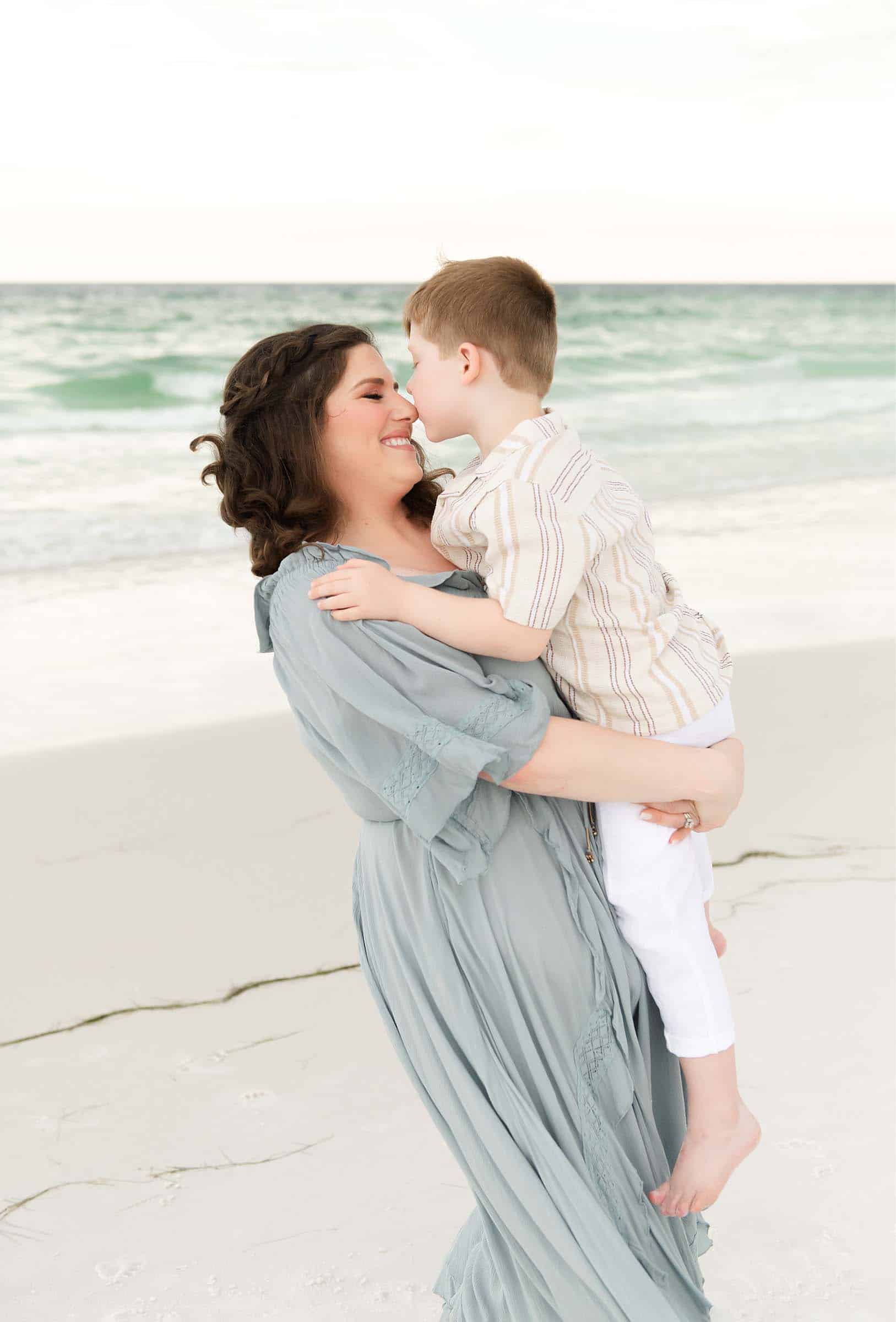 Woman in a light blue dress holds a young boy on the sand, both smiling for a Miramar beach family photographer.