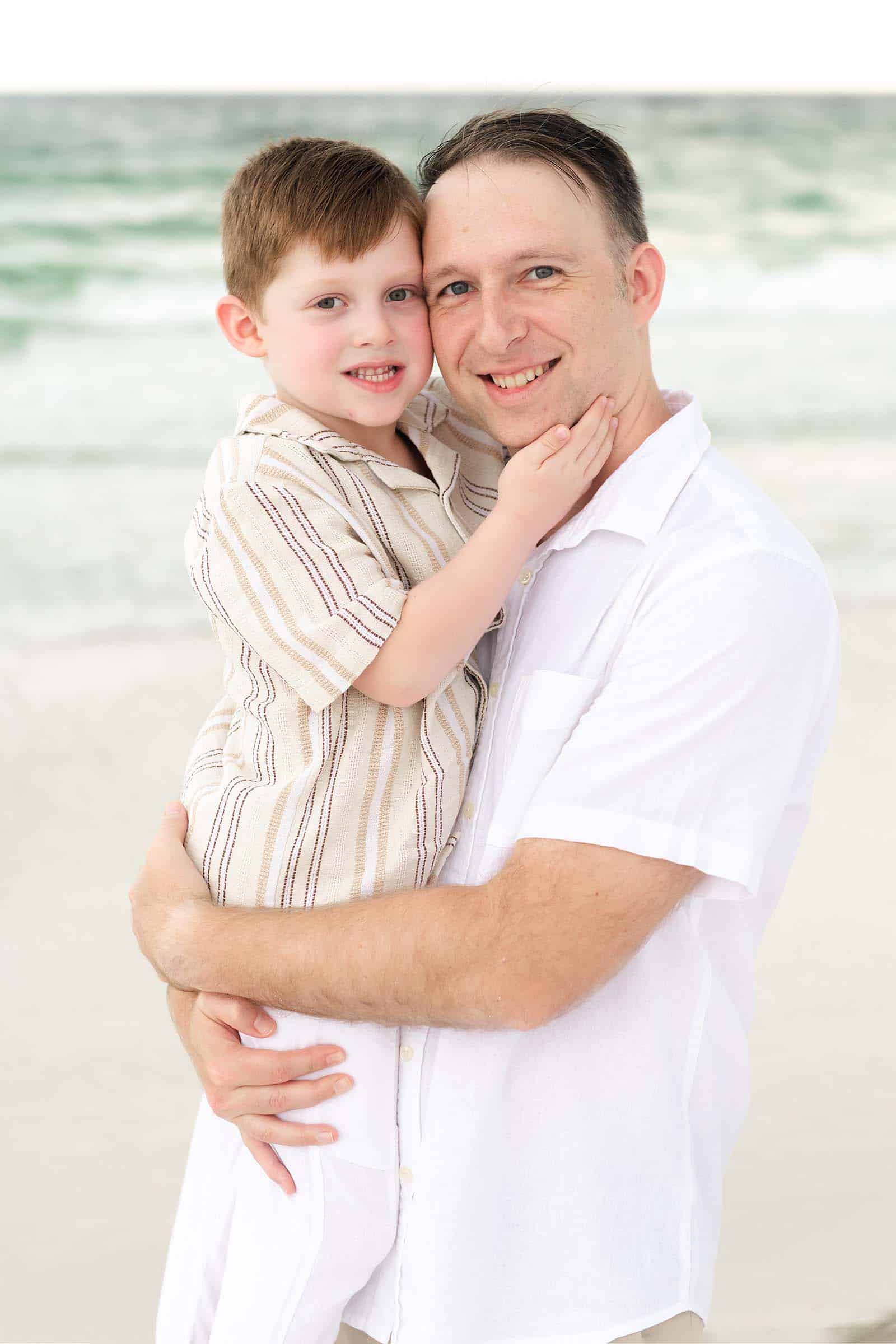 Father holding his son cheek to cheek on a Destin beach, both smiling at the camera, photographed by a Miramar Beach photographer