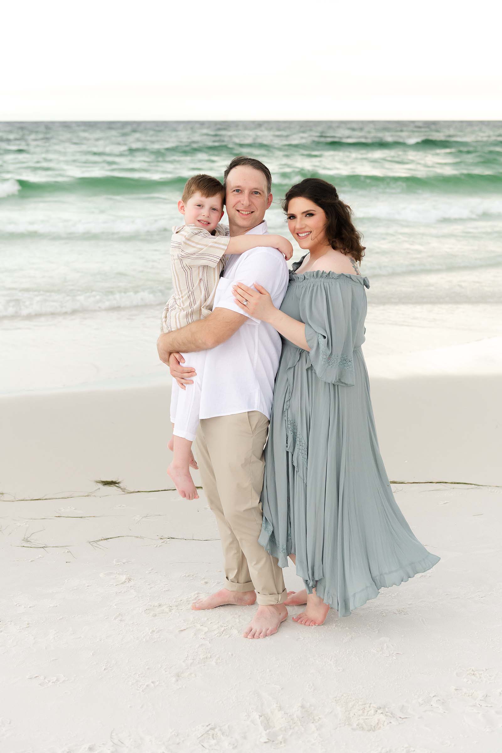 Family of three smiling together on a Destin beach, dad holding his son on his hip, photographed by a Miramar Beach family photographer