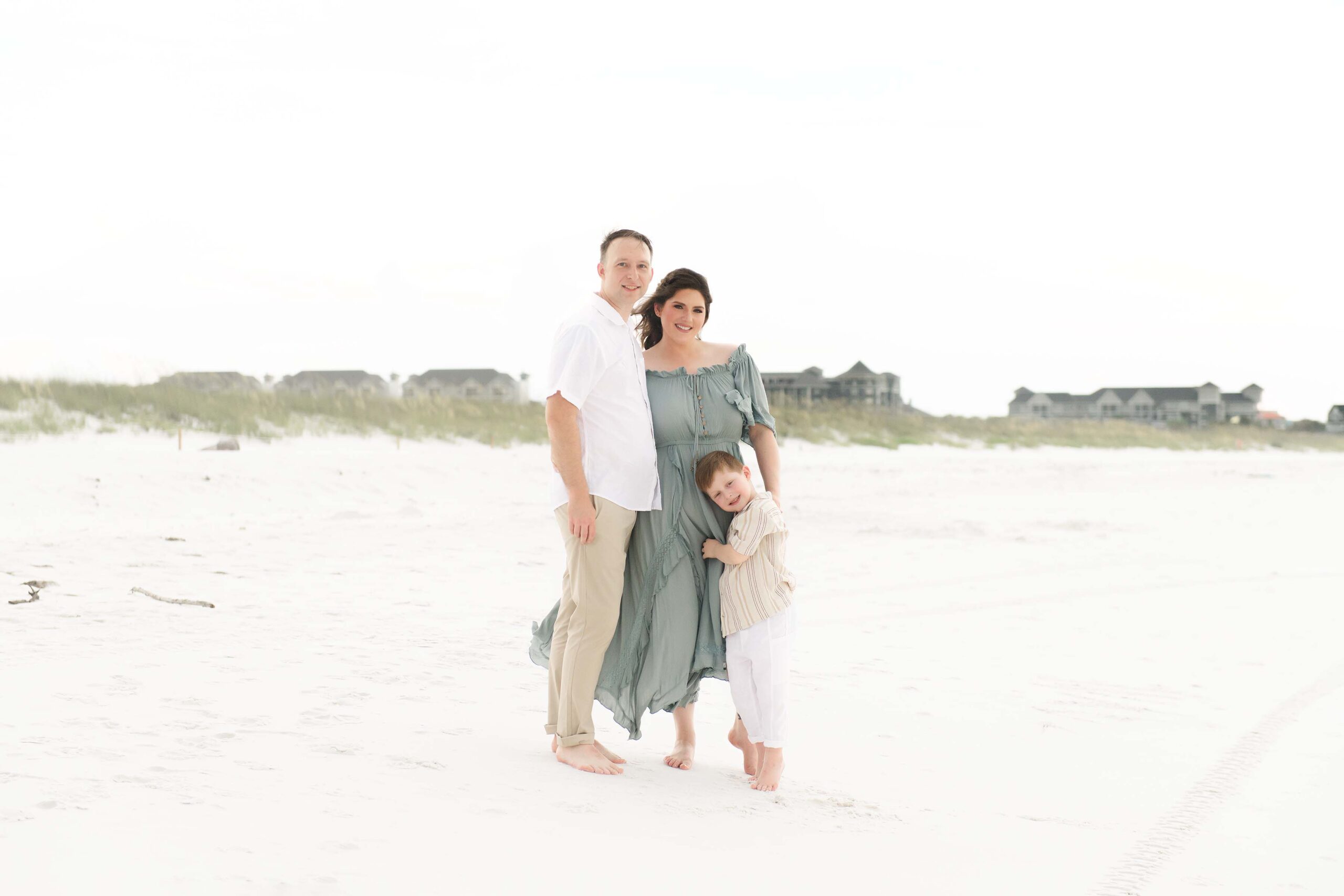 Family of three standing on white sand near the dunes, little boy hugging his mom, captured by photographers in Miramar Beach Florida