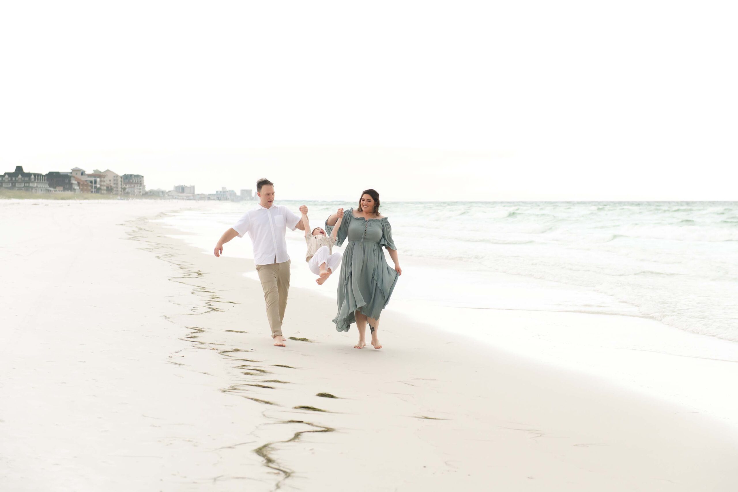 Parents swinging their little boy between them while walking on a Destin beach, all three laughing, photographed by a Miramar Beach family photographer