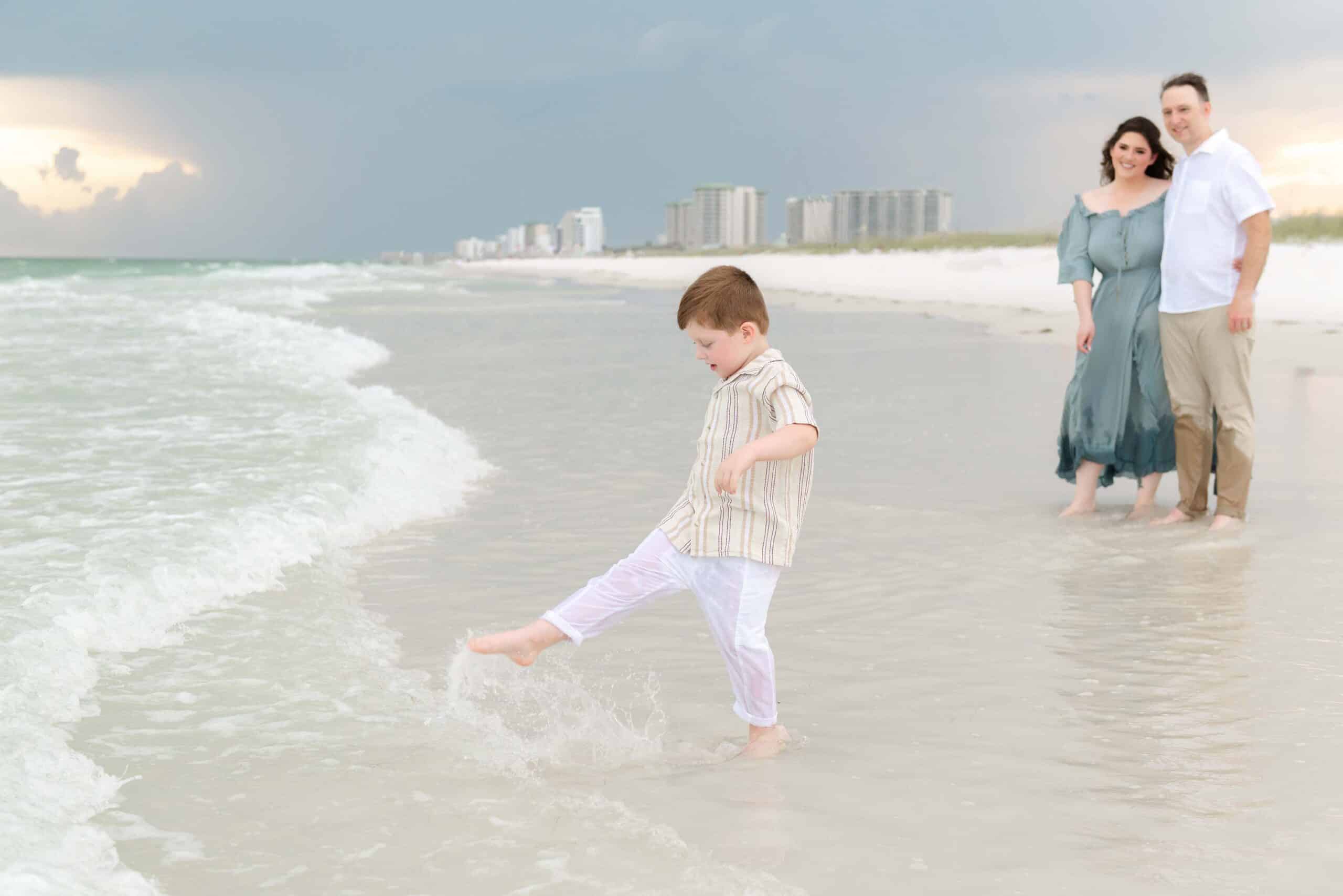 Little boy kicking water at the shoreline while his parents watch and smile behind him, captured by a Miramar Beach family photographer
