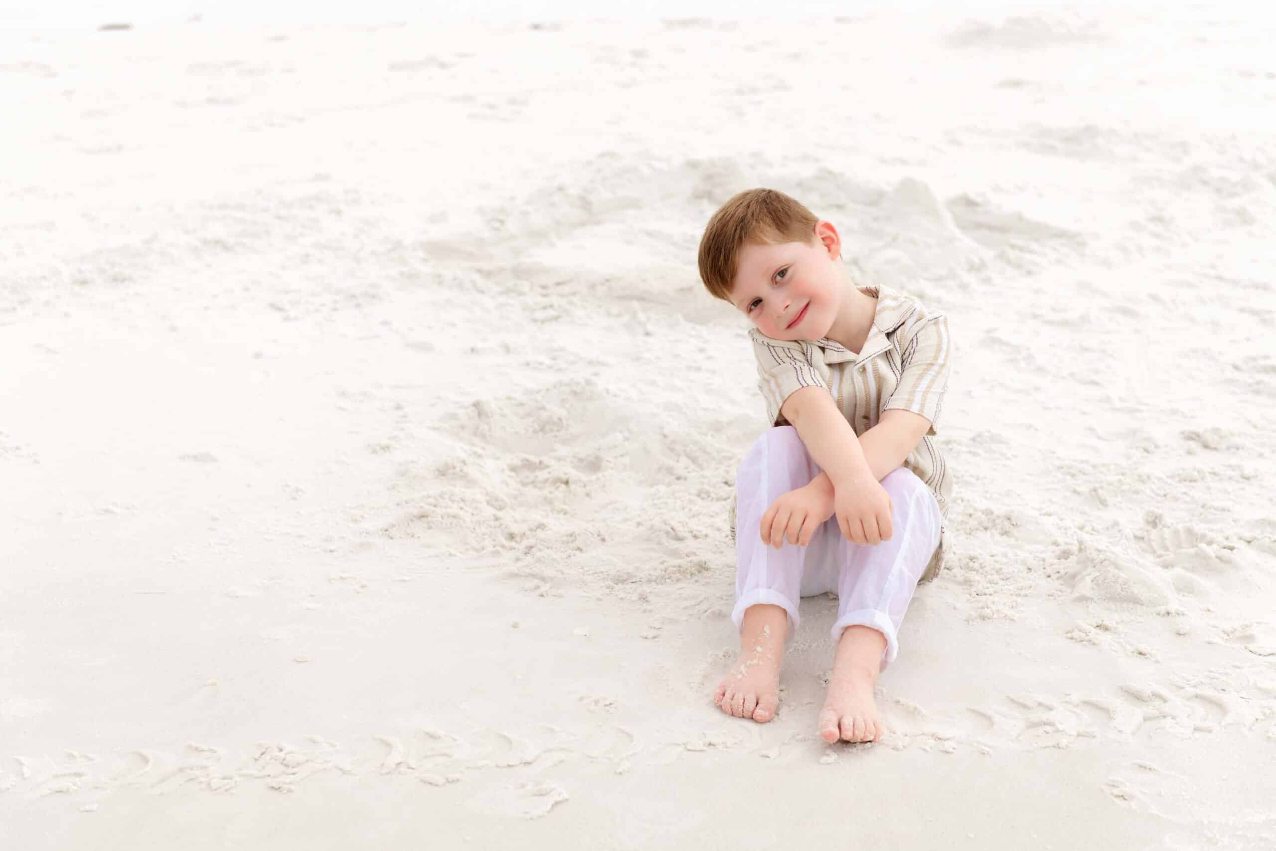 Little boy sitting on white sand smiling at the camera during a family vacation session near Miramar Beach Florida