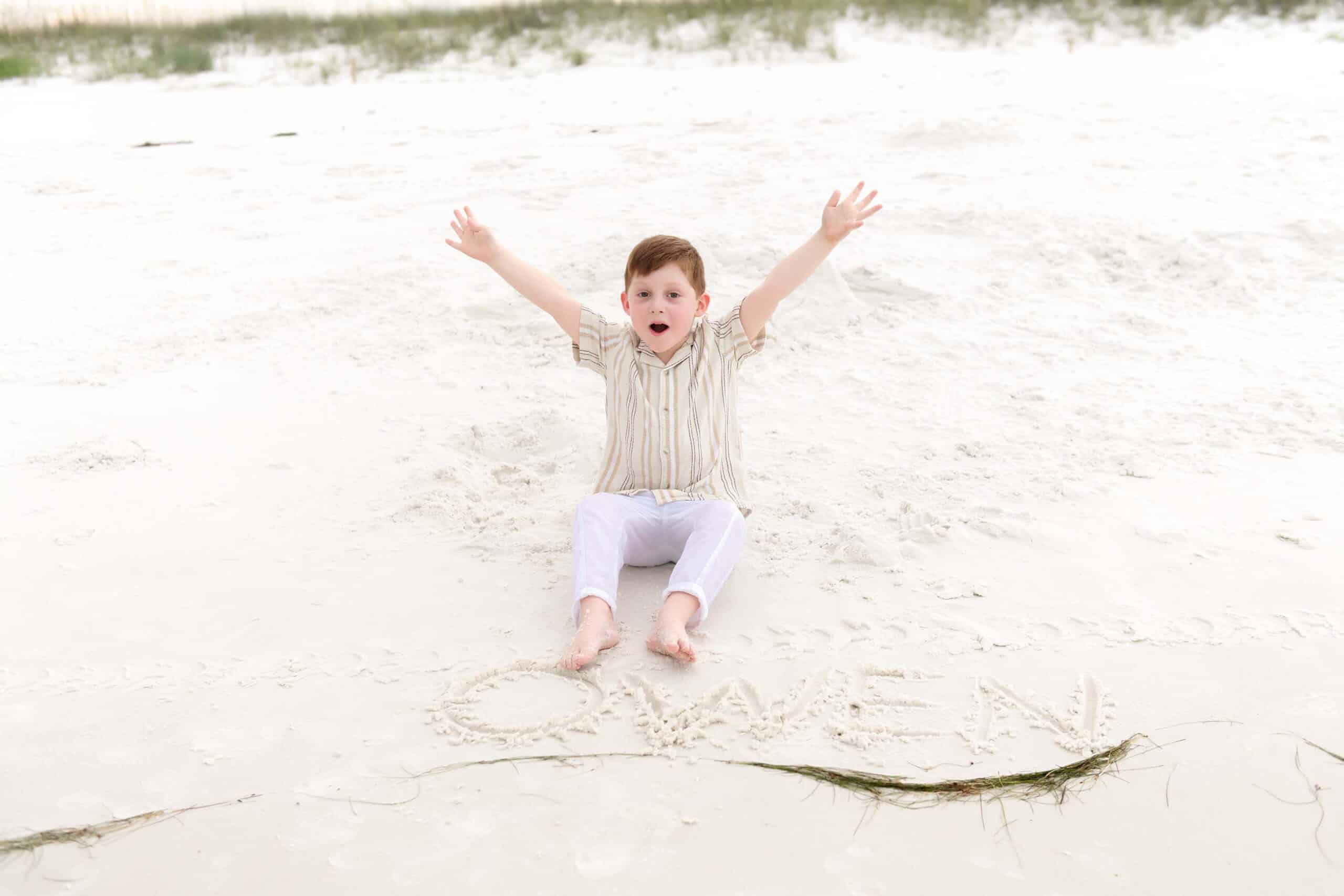 Little boy sitting in the sand with his arms raised in excitement during a family vacation session near Miramar Beach Florida