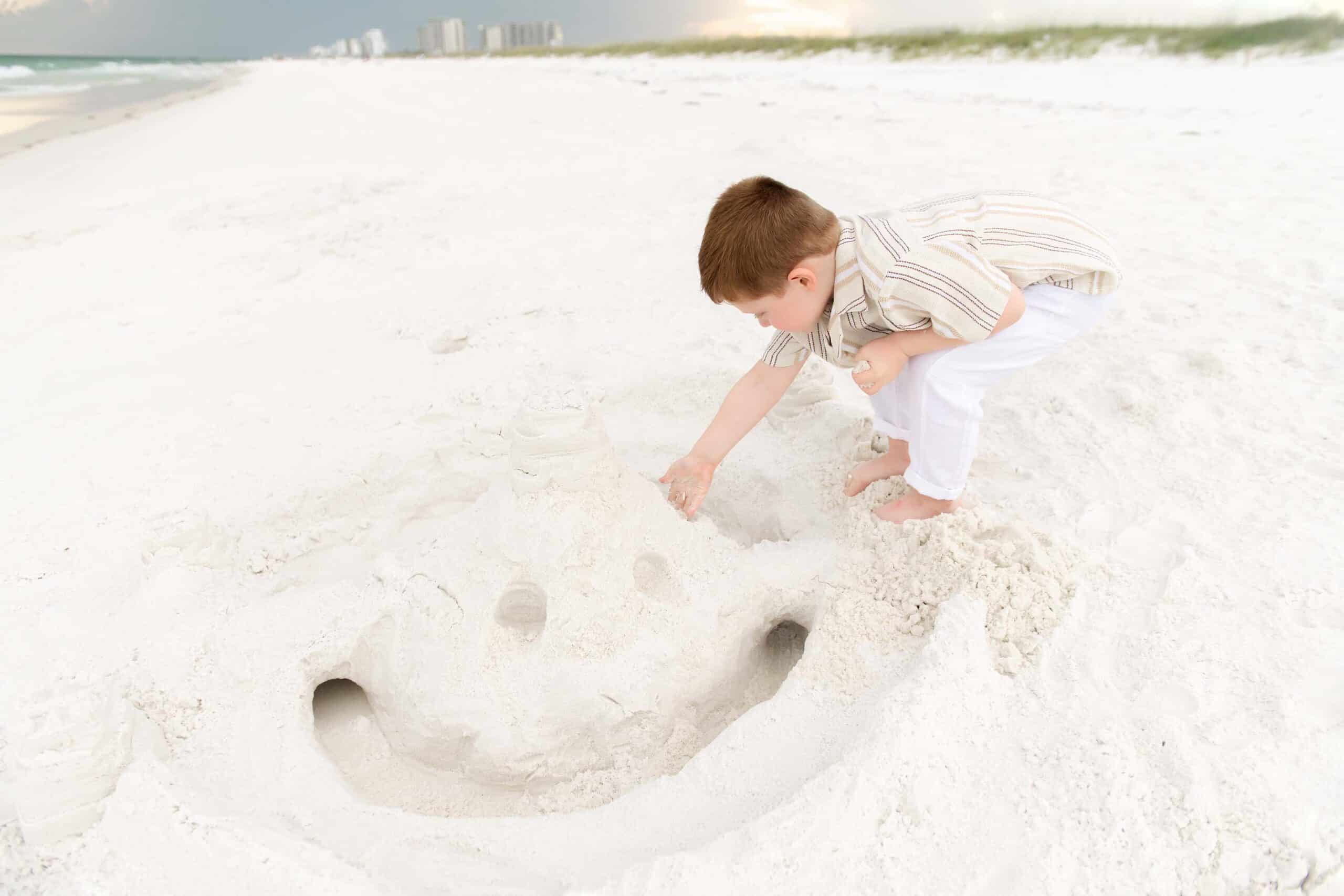 Little boy crouching over a sand smiley face he built on a Destin beach, captured by photographers in Miramar Beach Florida