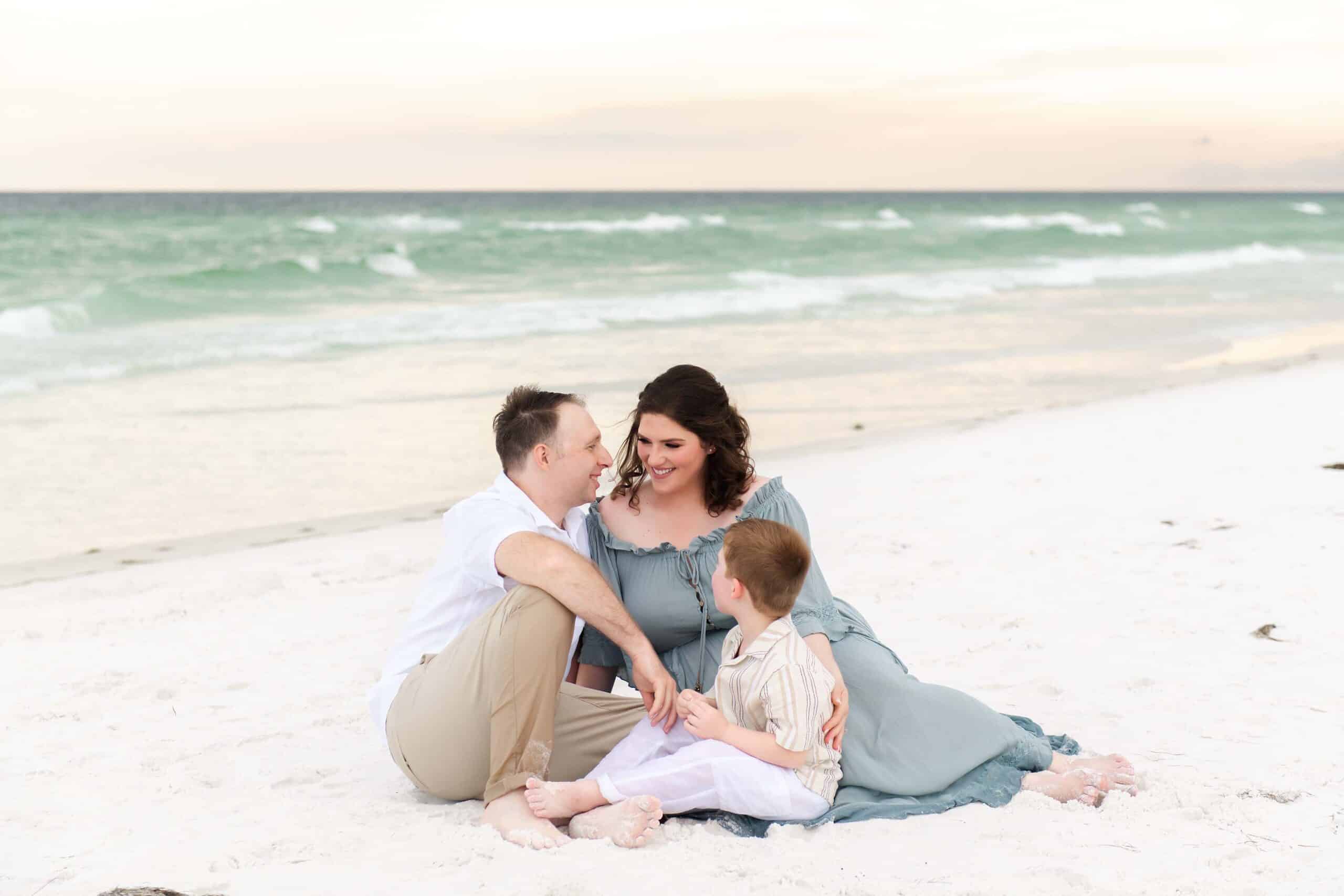 Family of three sitting together on a Destin beach at sunset, parents sharing a tender look with their little boy between them, photographed by a Miramar Beach photographer