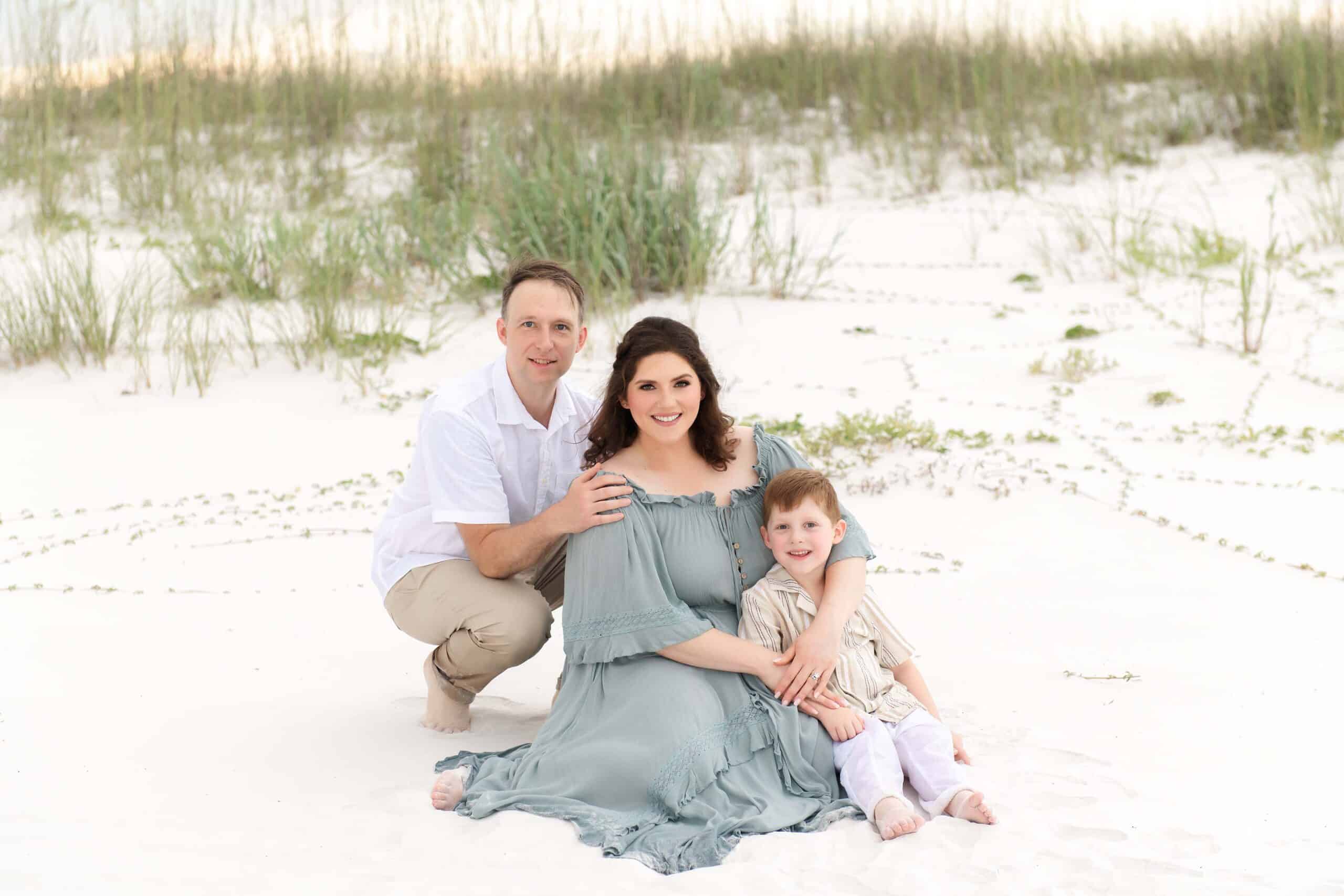 Family of three smiling at the camera in front of sea oats and dunes, photographed by photographers in Miramar Beach Florida