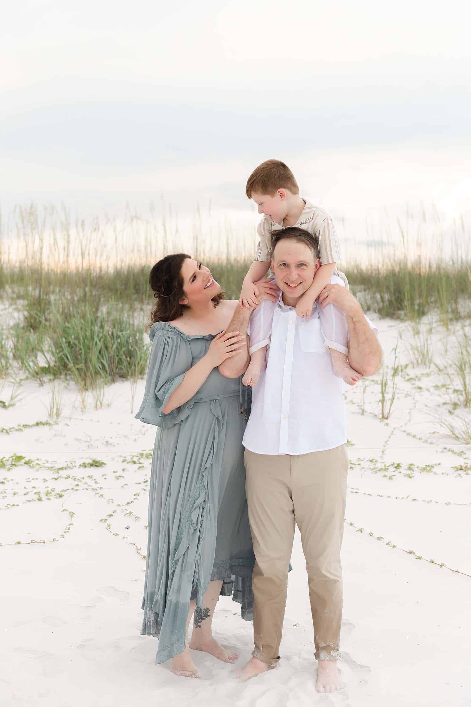 Little boy sitting on his dad's shoulders near the sea oats while mom laughs beside them, photographed by a Miramar Beach photographer