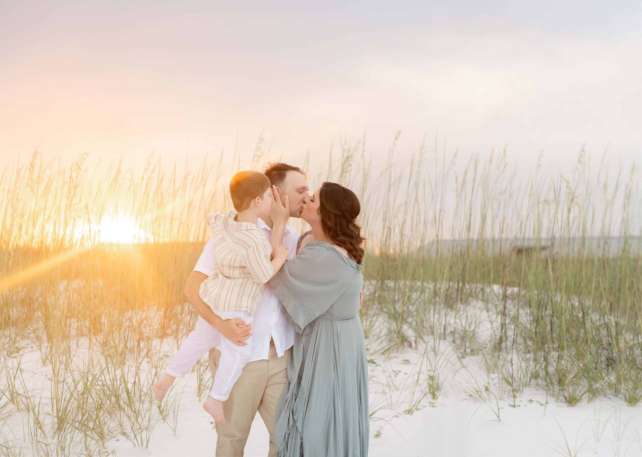 Family at golden hour sunset among the sea oats, parents sharing a kiss while dad holds their little boy, captured by a Miramar Beach family photographer