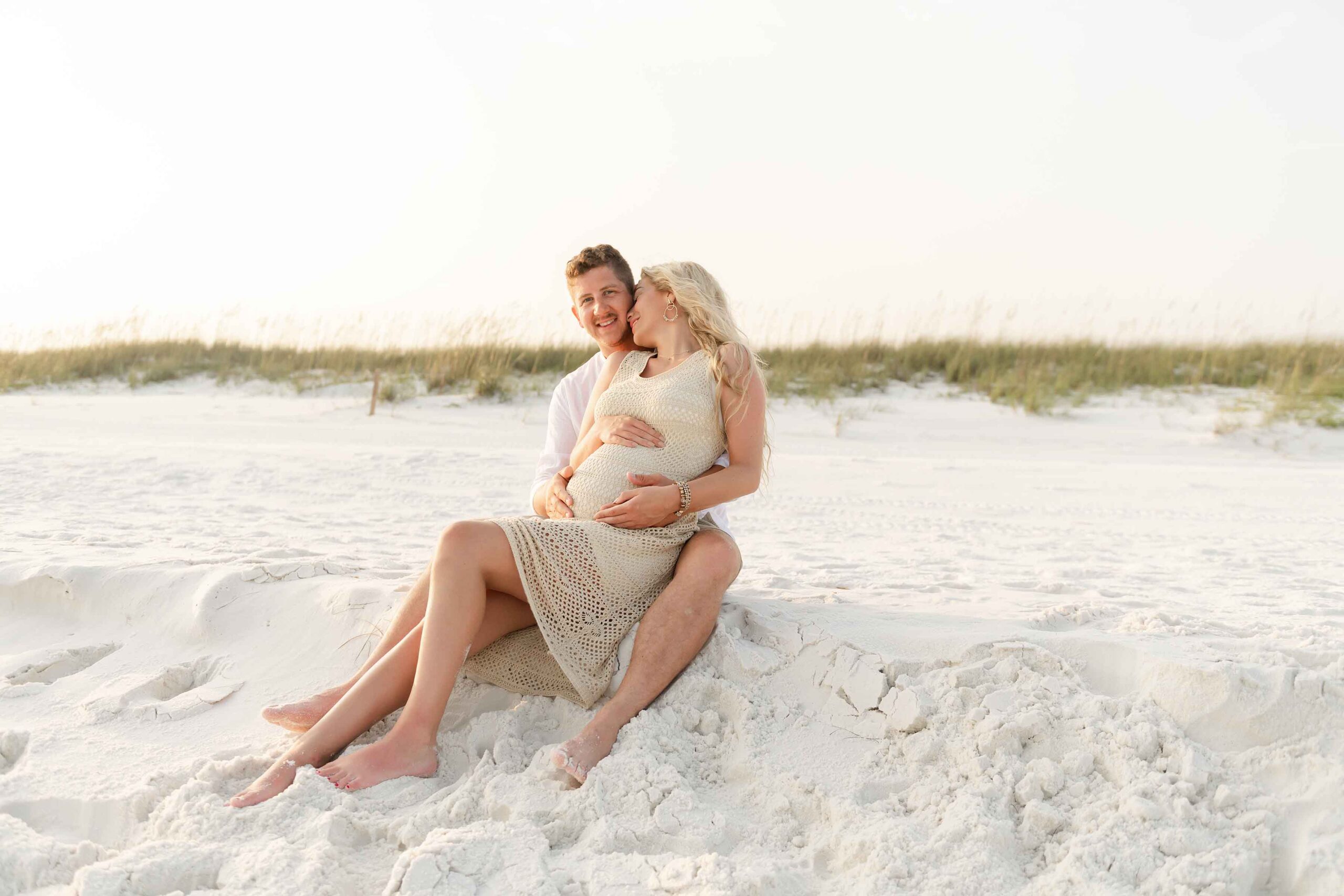 Pregnant couple sitting together on Destin beach sand, woman in cream knit dress, man in beige shorts and white shirt, sea oats visible