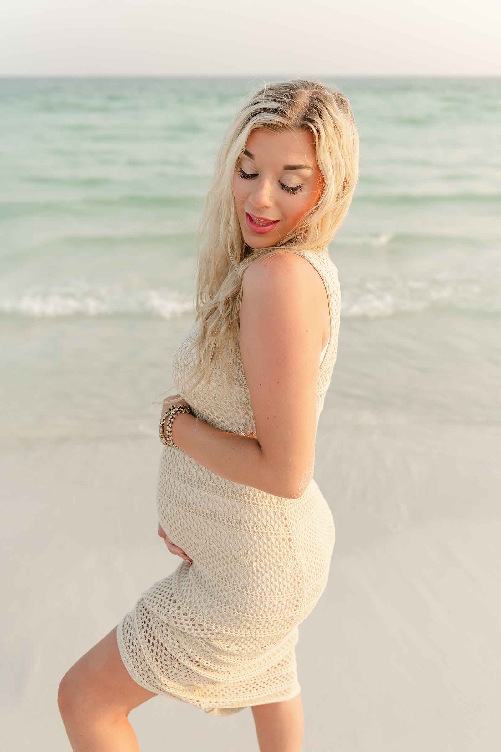Expecting mother in cream knit dress looking at camera during golden hour, wind in hair, Destin beach shoreline and dunes in soft focus