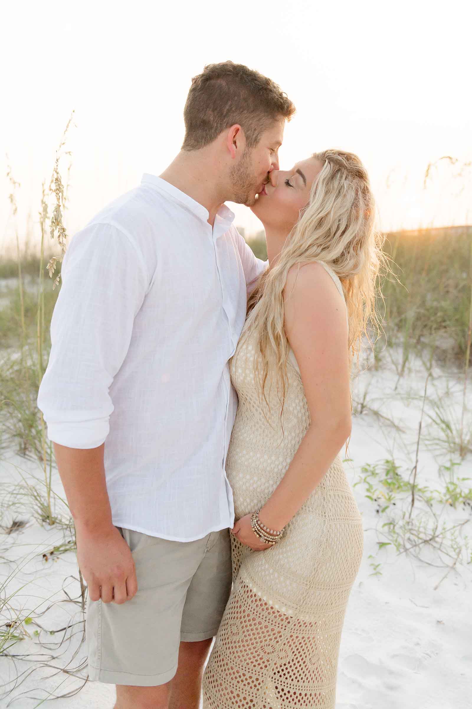Pregnant woman in cream knit dress side profile on Destin beach showing bump, golden hour backlighting, soft ocean and sky background