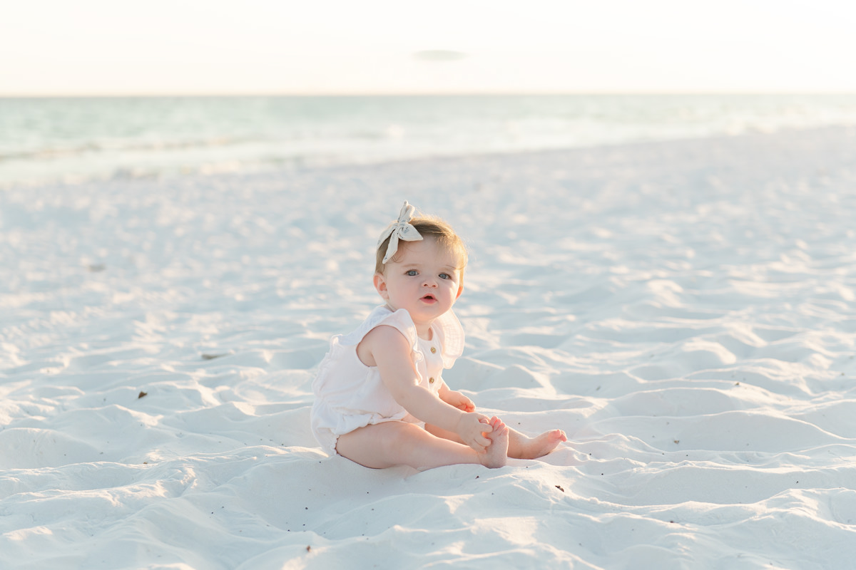 Baby in white sits on a sandy beach with ocean views&mdash;perfect for fall family photos in Destin.