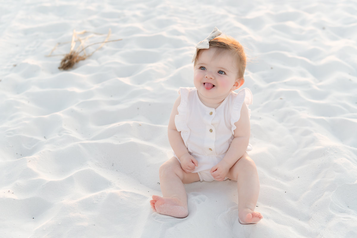 Baby sitting on white sand in romper and bow, sticking tongue out&mdash;perfect for fall family photos in Destin&rsquo;s charm.