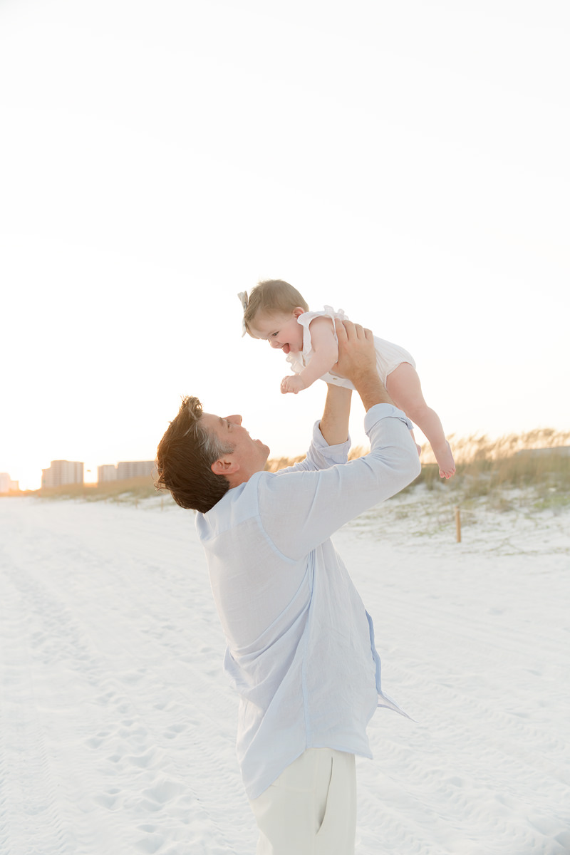 Man lifts a smiling baby in the air on a sandy beach at sunset&mdash;perfect for fall family photos in Destin.