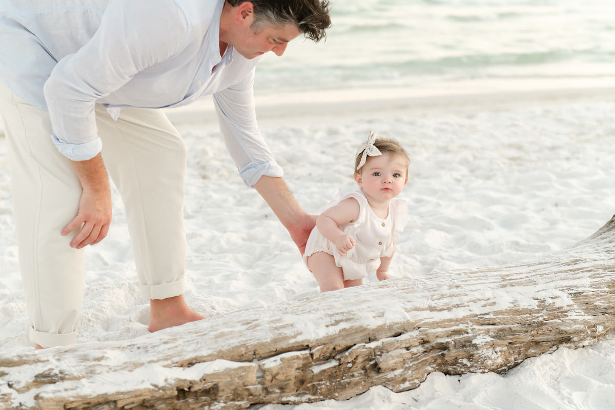 Man helps baby stand on sandy beach by driftwood, with ocean behind—perfect for fall family photos in Destin.