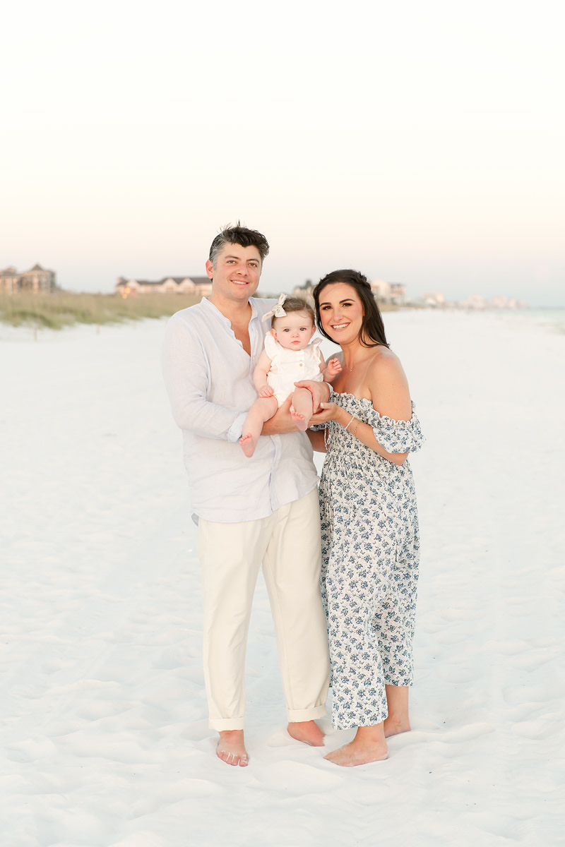 A couple stands barefoot on a sandy beach holding a baby&mdash;perfect for fall family photos in Destin with houses behind.