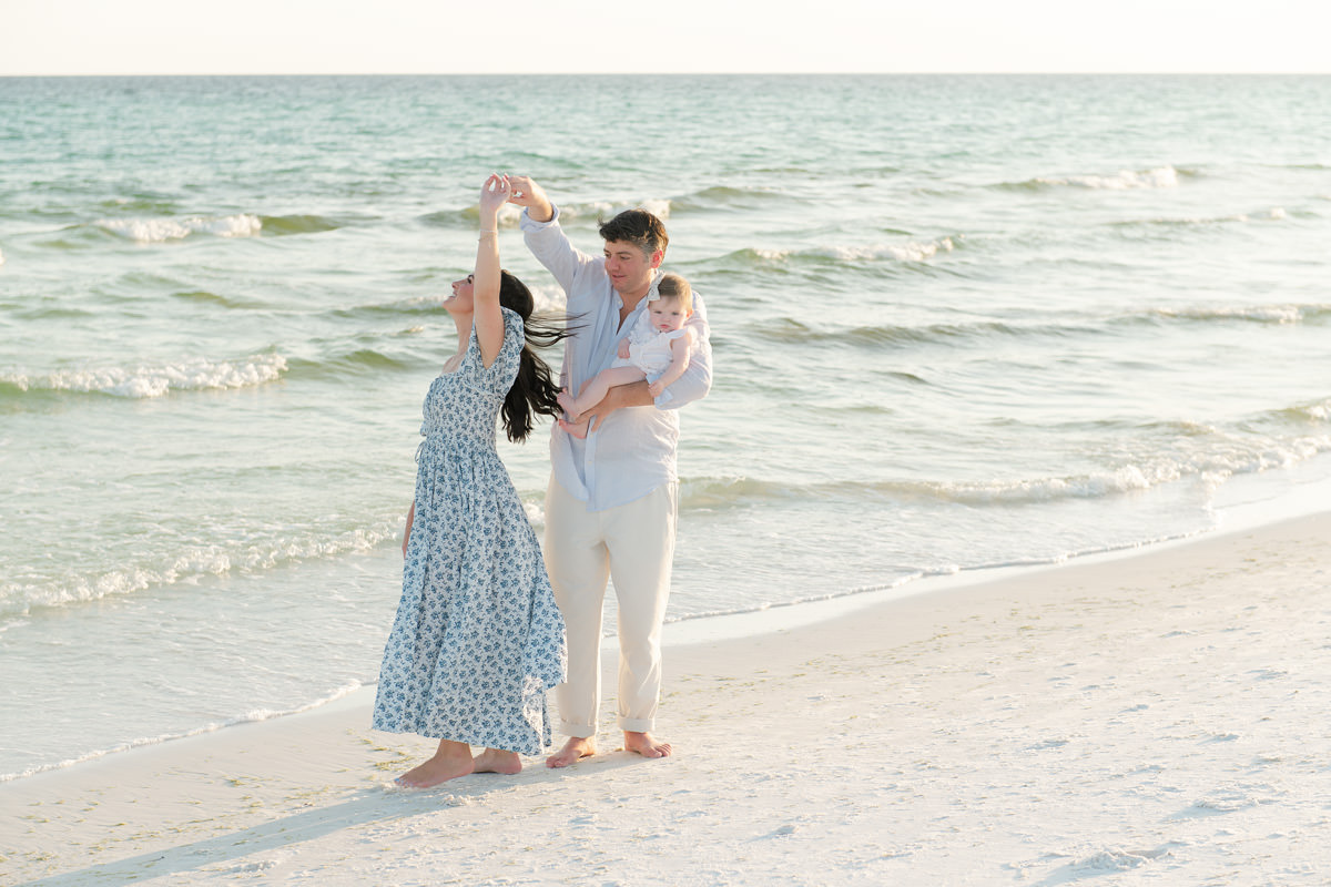 A man and woman dance on a sandy beach with a baby, capturing fall family photos in Destin by the ocean waves.