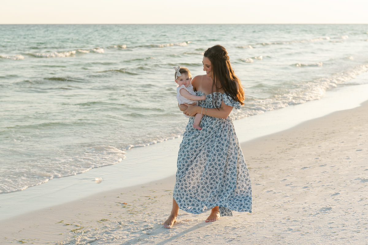 Woman in a blue floral dress holds a baby on a sandy Destin shoreline at sunset, perfect for fall family photos.