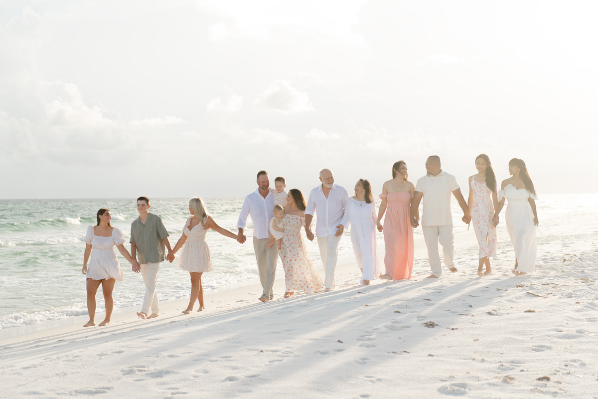 Extended family in coordinated cream and blush photo session outfits walking hand in hand on a white sand beach