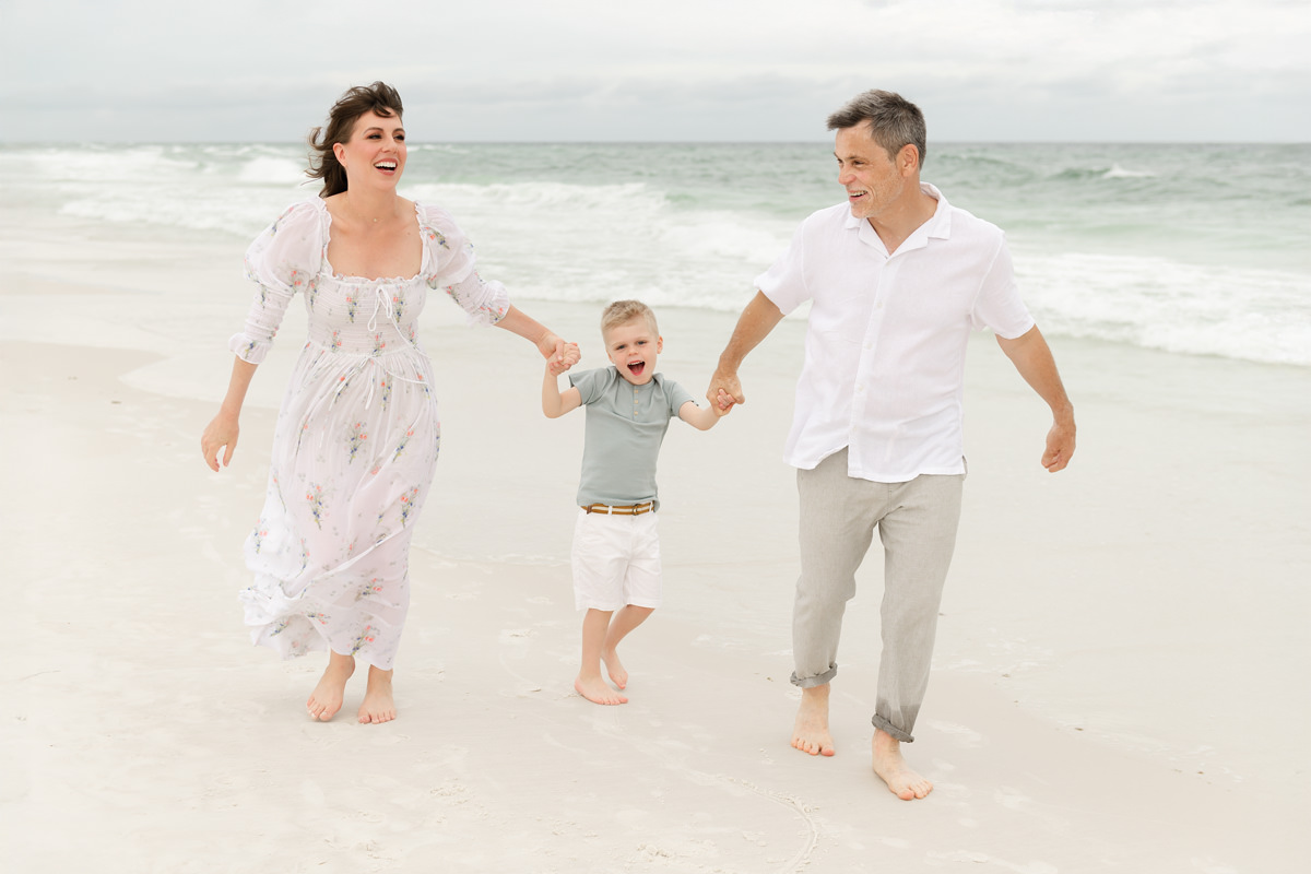 Family in white, sage, and floral session outfits laughing and holding hands while running on the beach