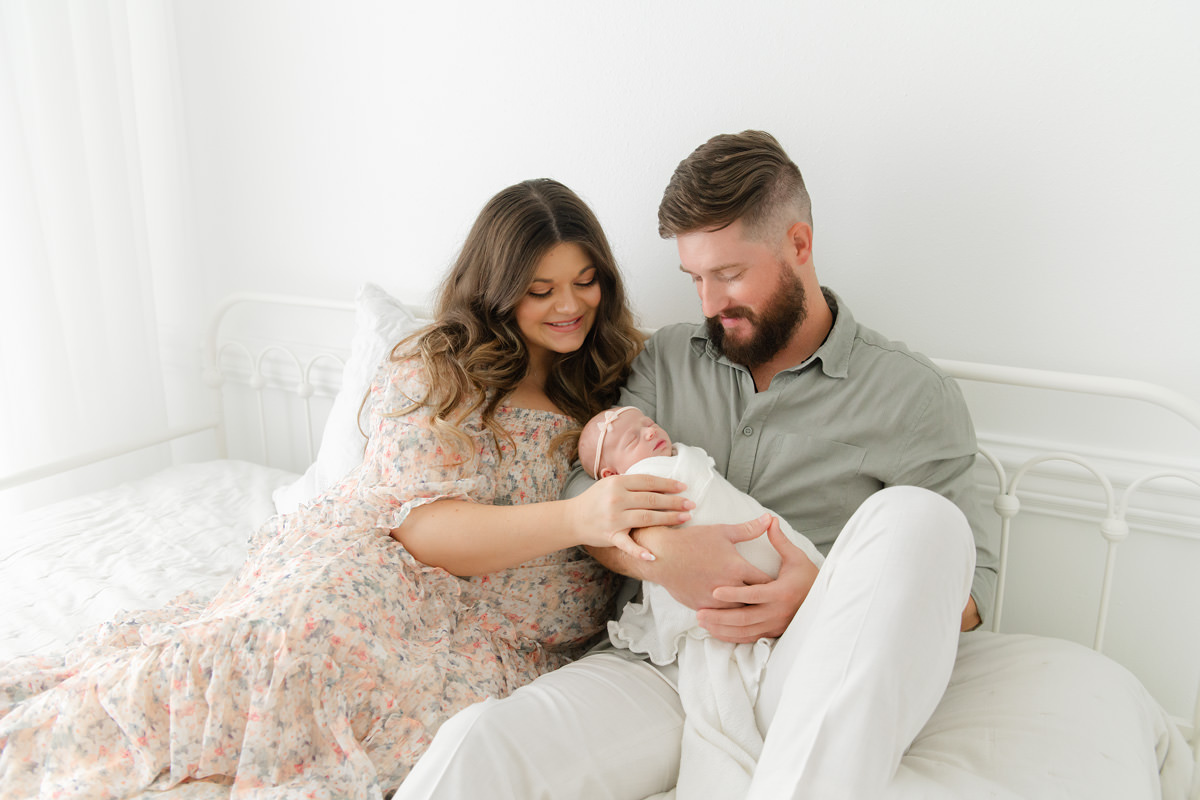 Parents in a peach floral dress and a sage green shirt holding their sleeping newborn on a white bed