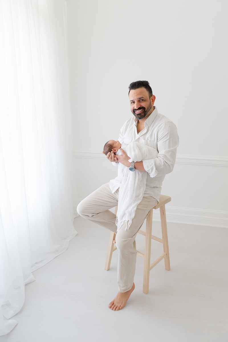 A man in a white shirt and light trousers holds a swaddled newborn baby during a Shalimar newborn photographer session.