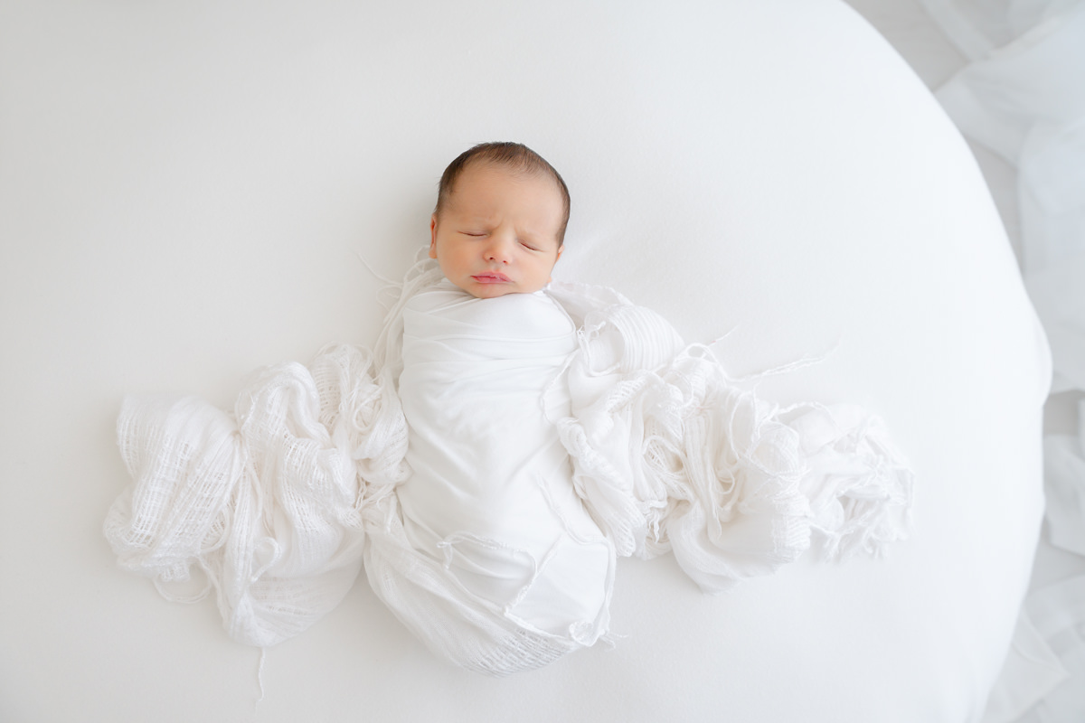 A newborn baby swaddled in white fabric sleeping on a white surface, captured by a Shalimar newborn photographer.