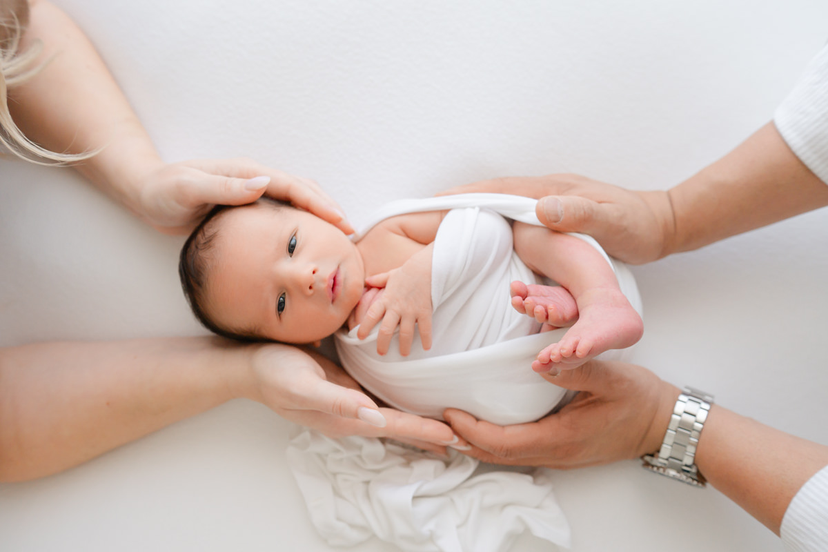 A newborn swaddled in white cloth being held by two adults, captured by a Shalimar newborn photographer.