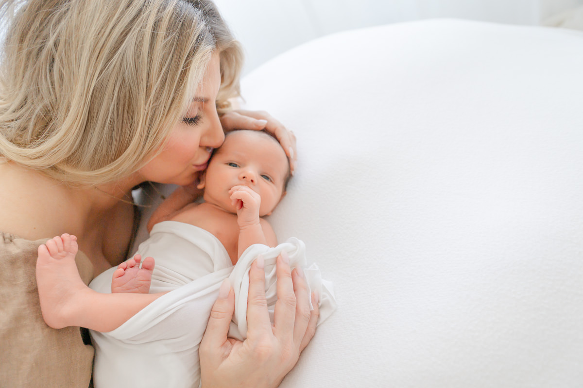 A Shalimar newborn photographer captures a woman kissing the forehead of a baby wrapped in a white cloth.