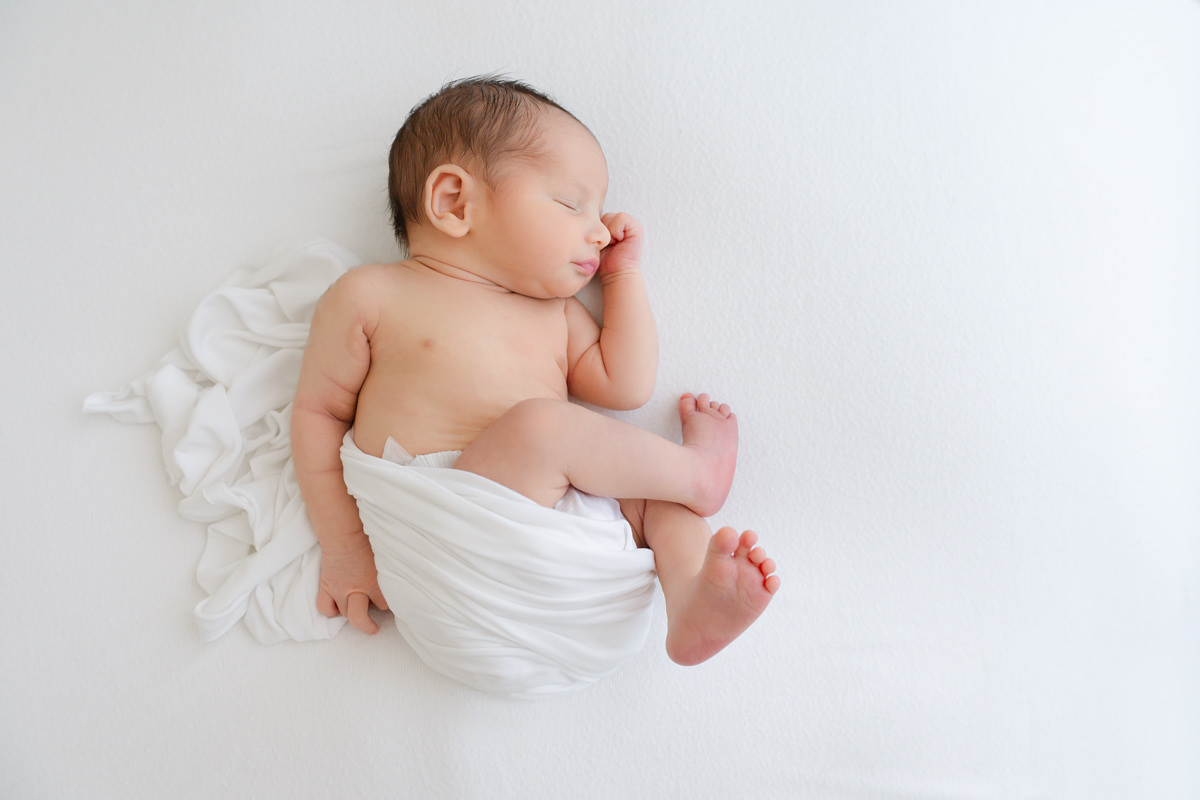 A newborn baby sleeping on a white surface, partially wrapped in a white cloth, captured by a Shalimar newborn photographer.