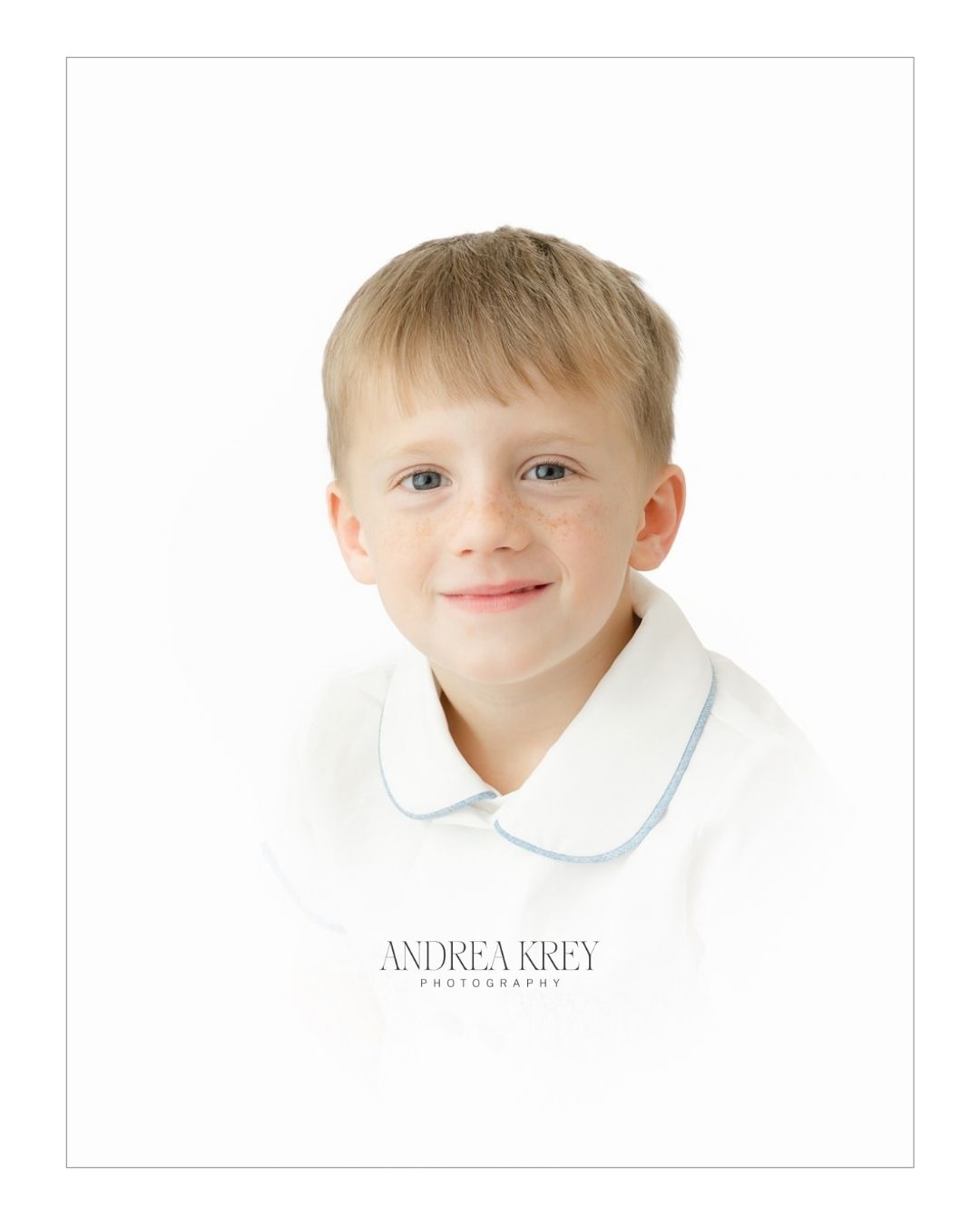 Smiling boy with freckles in a white collared shirt with blue trim on a white vignette backdrop